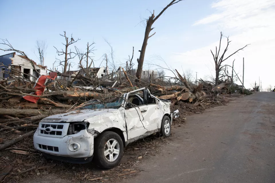 Heavily damaged white small SUV parked on street with heavy tornado damage and leveled houses in Mayfield, Kentucky.